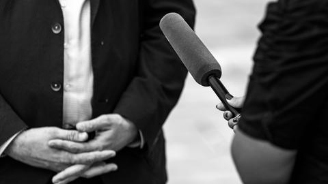 Schwarz-Weiß Bild (Ausschnitt) von einer Person, die eine andere interviewt, man sieht ein Mikrofon und gefaltete Hände.