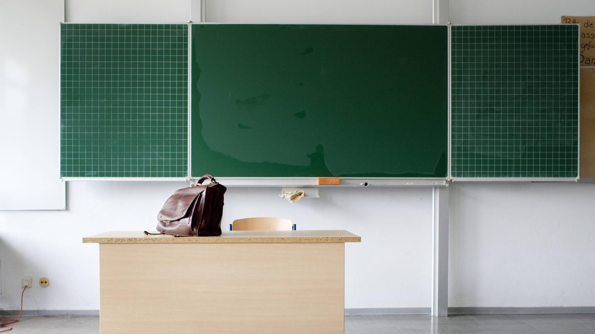 Eine Ledertasche auf einem leeren Lehrerpult. Dahinter eine Schultafel. 