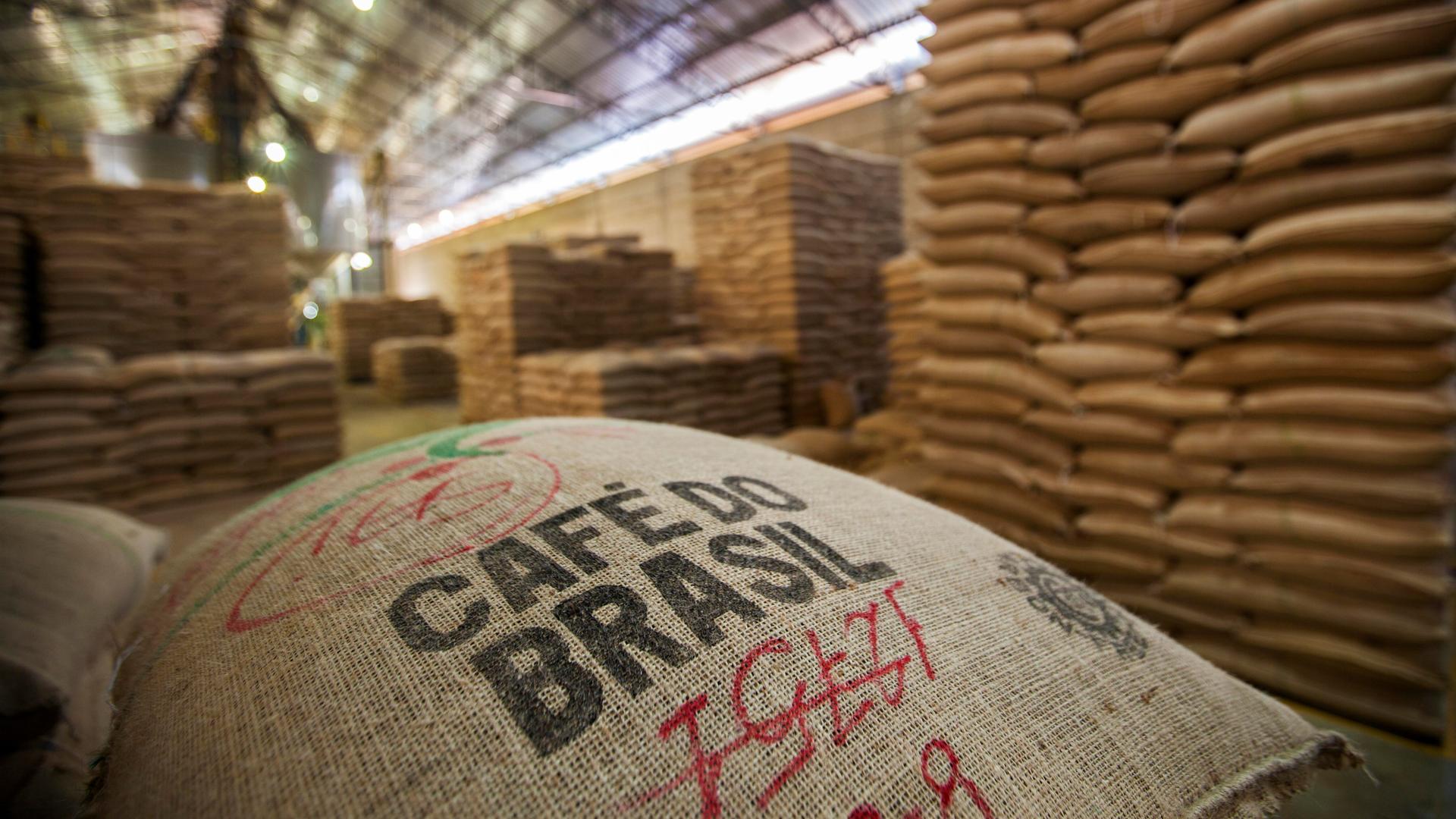 Im Vordergrund liegt ein Kaffeesack mit der Aufschrift "Café do Brasil" – zu deutsch "Kaffee aus Brasilien". Im Hintergrund sind in der Lagerhalle weitere Säcke gestapelt.