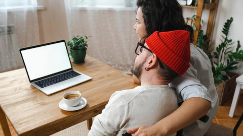 Zwei Menschen schauen auf einen Laptopbildschirm. Sie sind von hinten zu sehen. Die Frau rechts, mit dunklen Locken, legt ihren Arm um den Mann links mit roter Mütze. Er sitzt im Rollstuhl. 