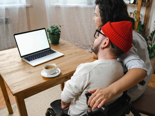 Zwei Menschen schauen auf einen Laptopbildschirm. Sie sind von hinten zu sehen. Die Frau rechts, mit dunklen Locken, legt ihren Arm um den Mann links mit roter Mütze. Er sitzt im Rollstuhl. 