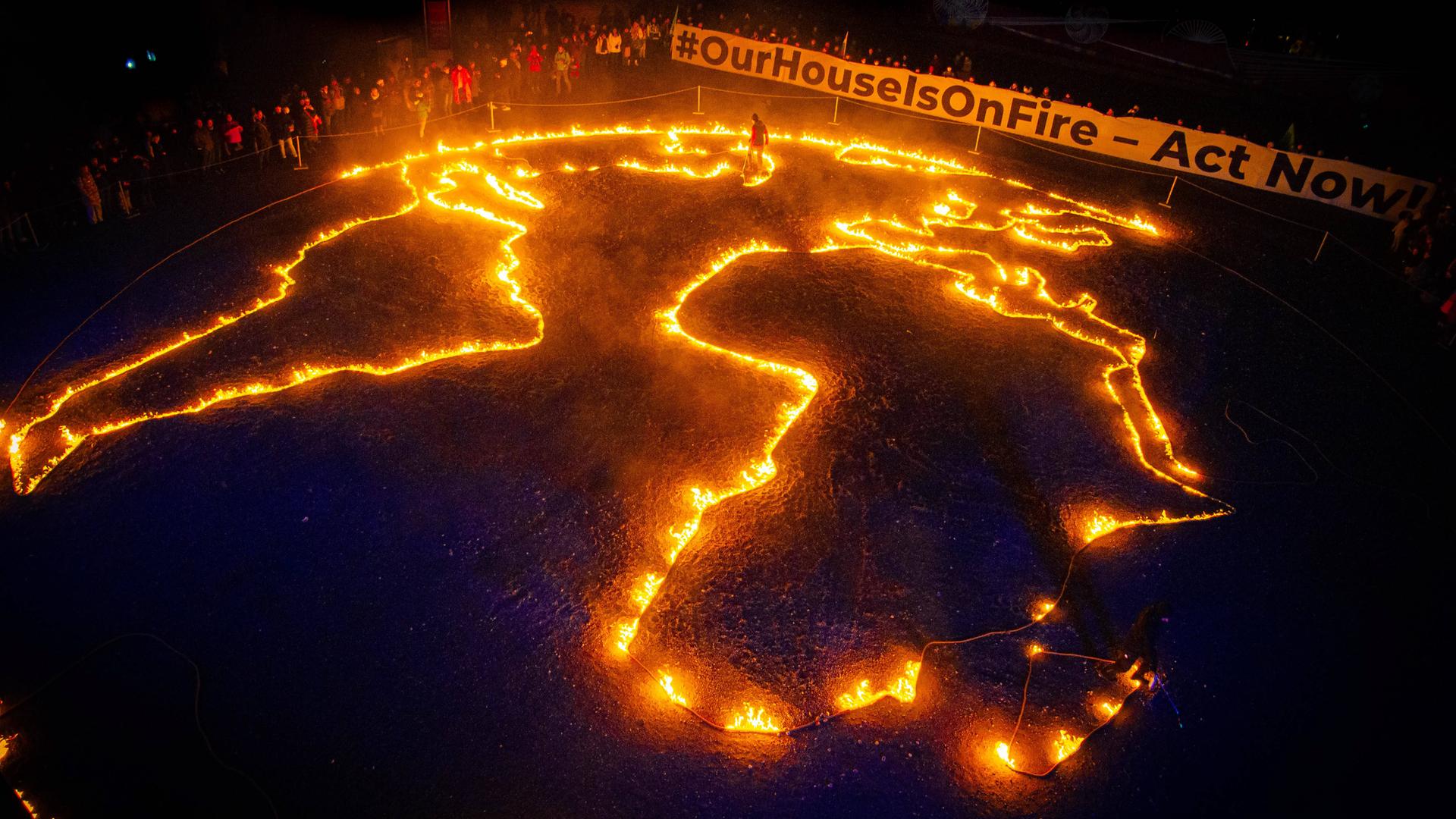 Ein auf den Boden gelegter Umriss der Erde von 30 Metern wird in Brand gesetzt. Darüber hält eine Menschenmege ein Spruchband auf dem steht "#OurHouseIsOnFire - Act Now"