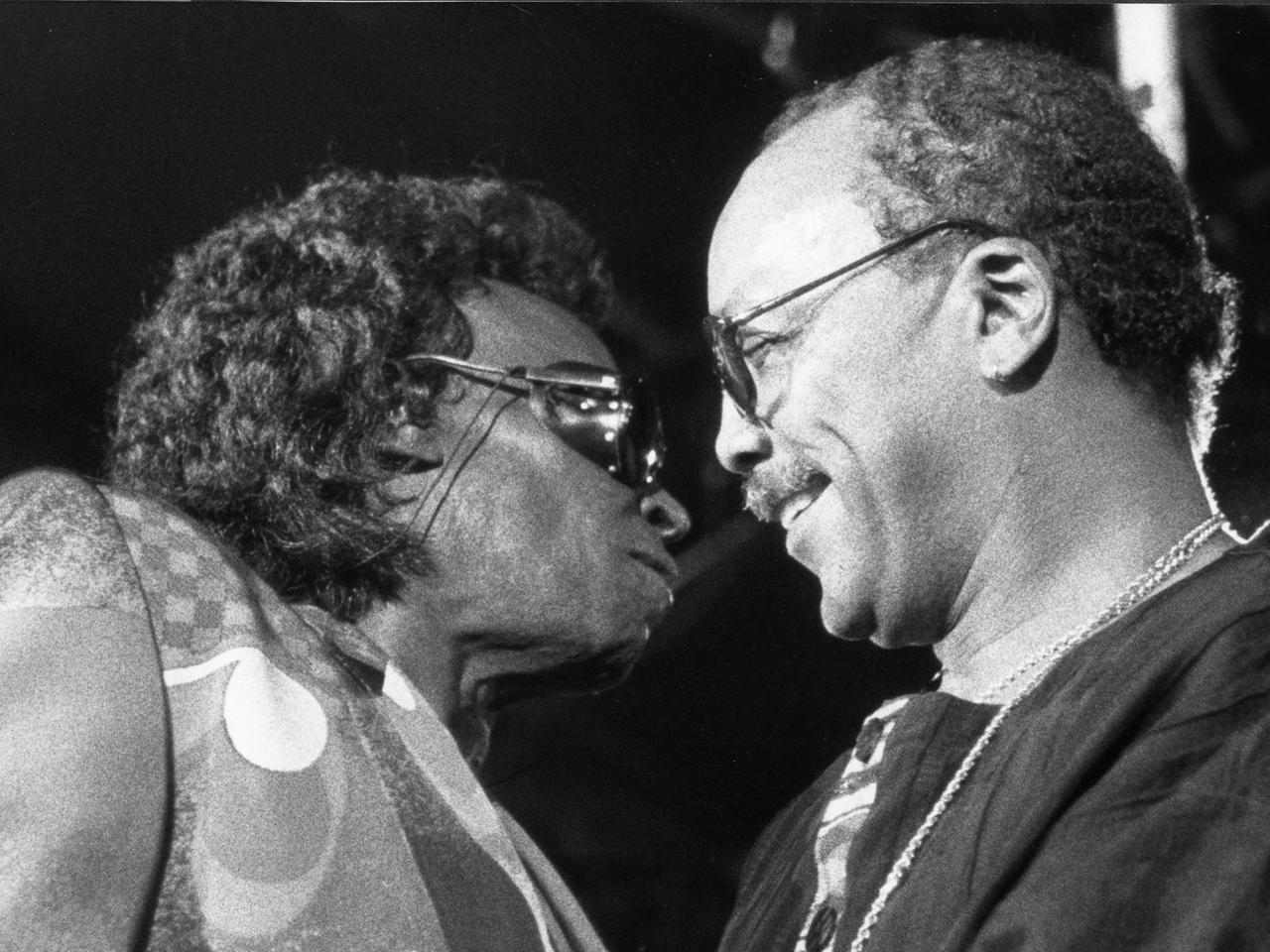 Miles Davis in hellem Anzug, mit vollem Haar und Sonnenbrille, steht ganz dicht vor Quincy Jones, der ebenfalls eine Brille trägt und schüttereres Haar hat.Jones lächelt. Das Bild zeigt beide Musiker im Profil.