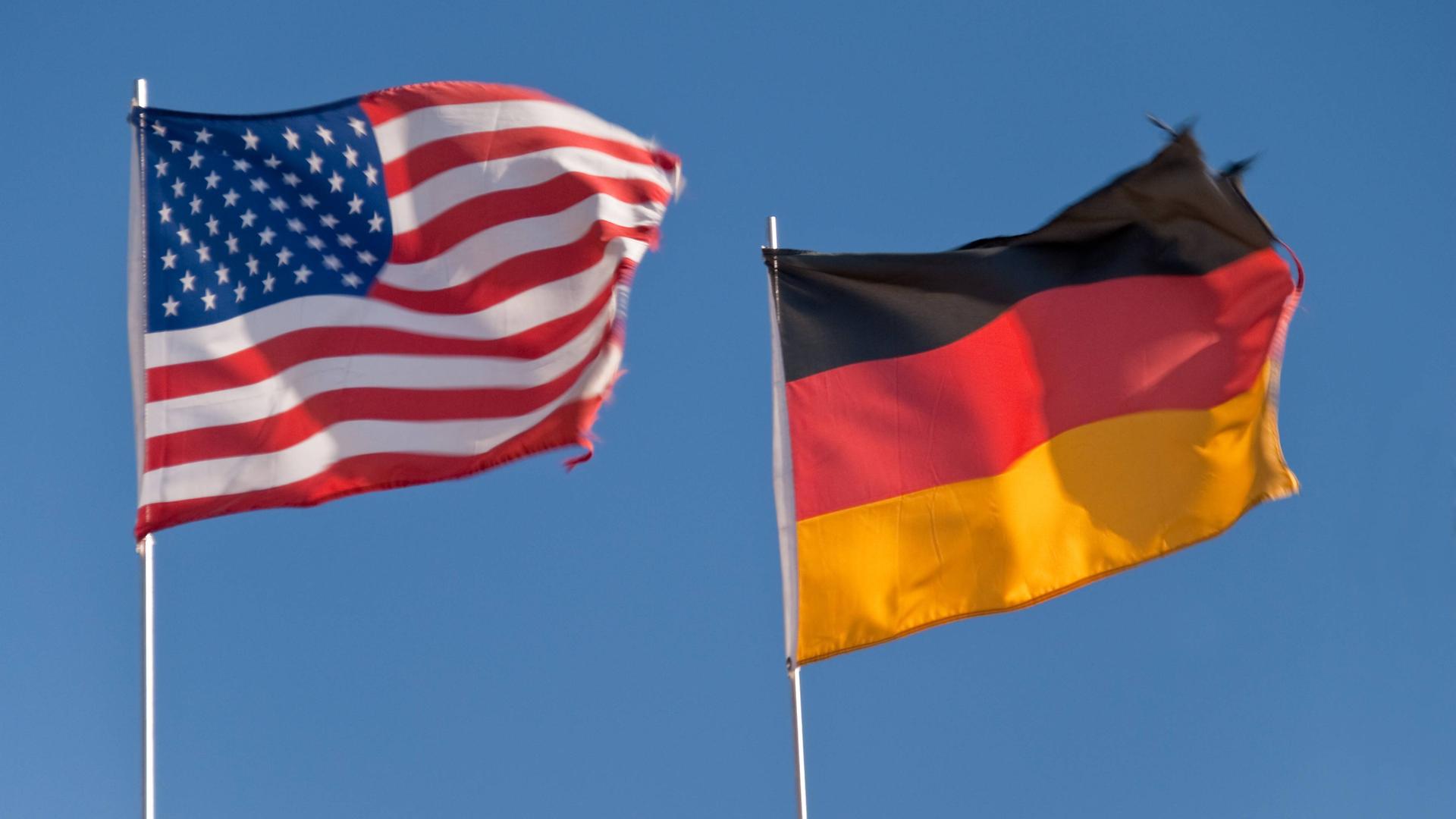 Eine US-amerikanische und ein deutsche Flagge flattern im Wind, dahinter blauer Himmel