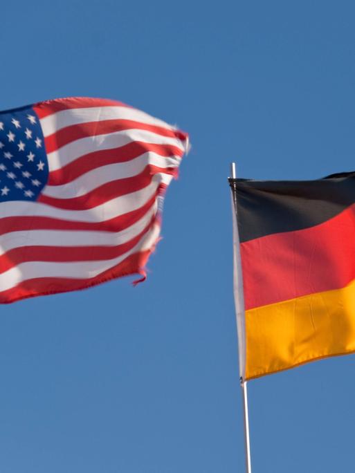 Eine US-amerikanische und ein deutsche Flagge flattern im Wind, dahinter blauer Himmel