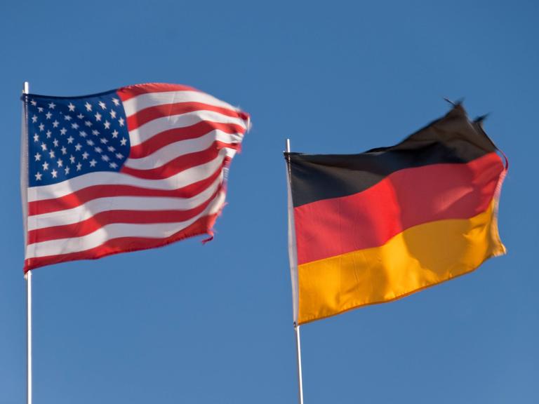 Eine US-amerikanische und ein deutsche Flagge flattern im Wind, dahinter blauer Himmel