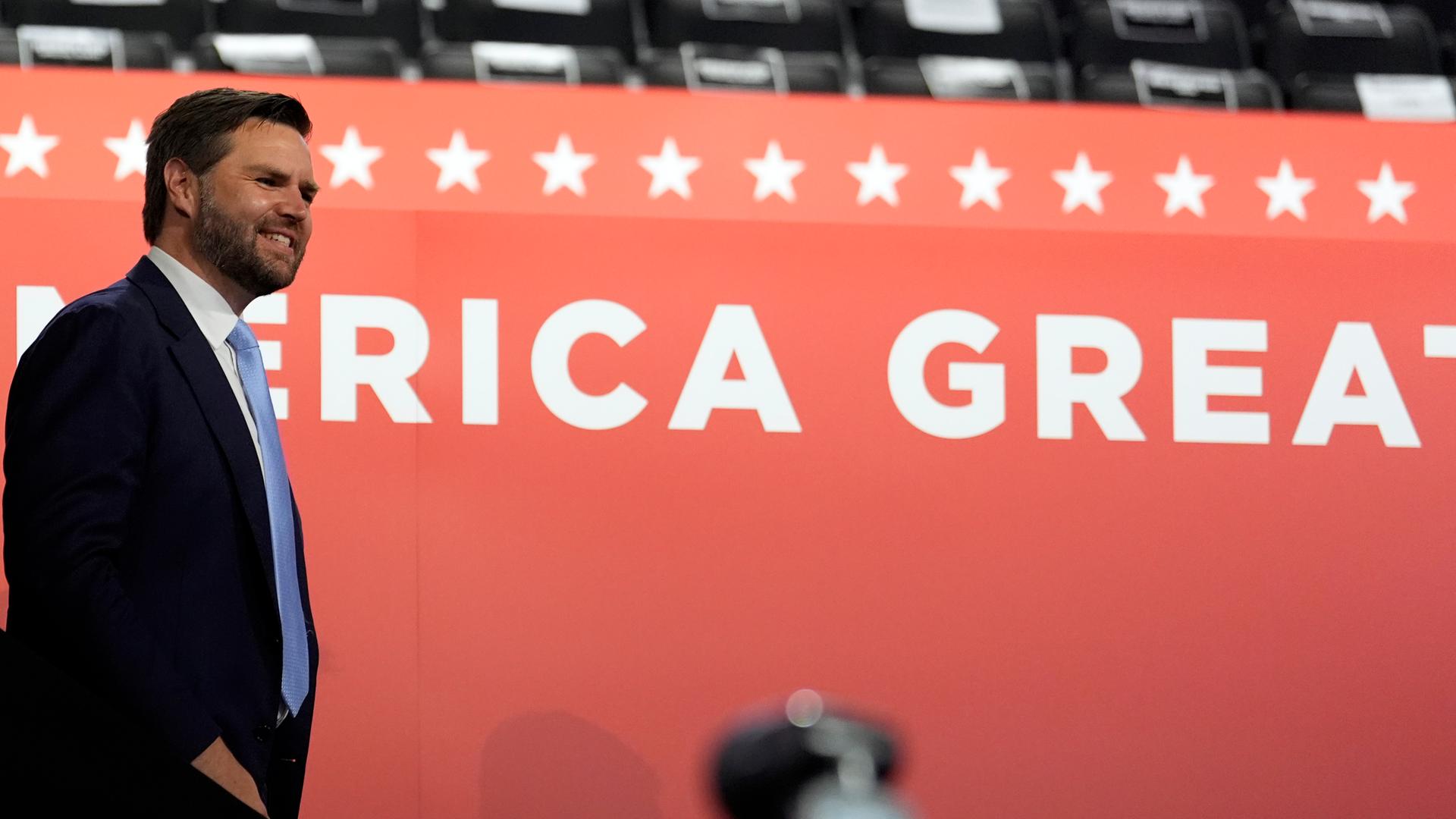 Trumps Vize J.D Vance steht vor einer roten Wand mit dem Schriftzug "Make America great again".