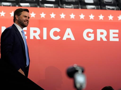 Trumps Vize J. D. Vance steht vor einer roten Wand mit dem Schriftzug "Make America great again".