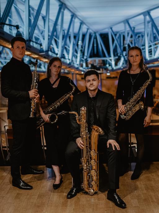 Zwei junge Frauen und zwei junge Männer stehen mit ihren unterschiedlich großen Saxophonen in einem modernen Konzertsaal mit Stahlträgerkonstruktionen.