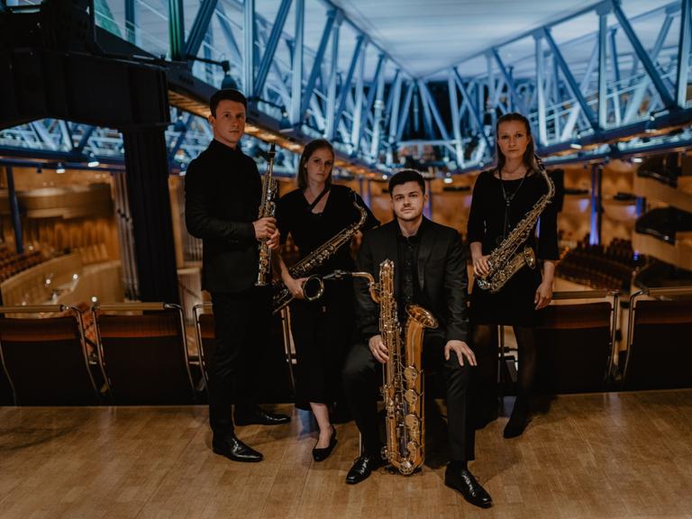 Zwei junge Frauen und zwei junge Männer stehen mit ihren unterschiedlich großen Saxophonen in einem modernen Konzertsaal mit Stahlträgerkonstruktionen.
