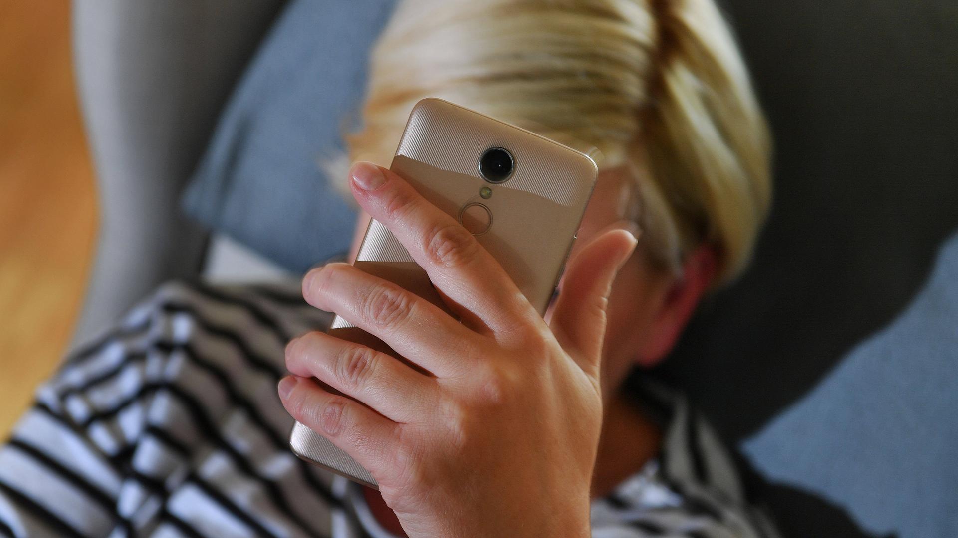 Eine Person hält ein Smartphone in der Hand. Das Gesicht ist durch das Smartphone verdeckt.
