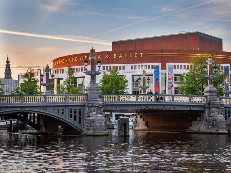 Außenaufnahme des Opernhauses in Amsterdam. Davor eine Brücke und eine Gracht. Dahinter Abendhimmel.