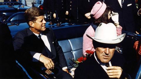 John F. Kennedy sitzt lachend in einer Limousine, neben ihm befindet sich seine Frau Jackie Kennedy.