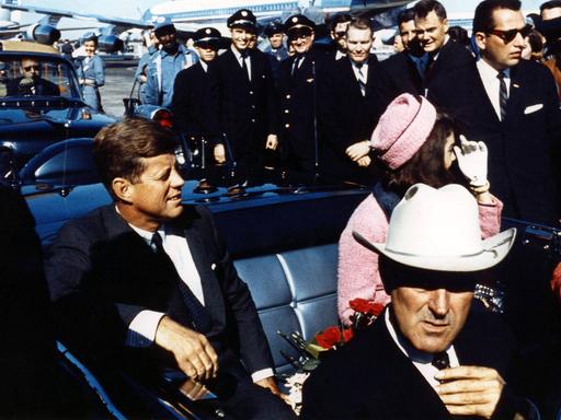 John F. Kennedy sitzt lachend in einer Limousine, neben ihm befindet sich seine Frau Jackie Kennedy.