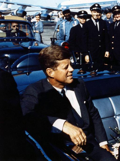 John F. Kennedy sitzt lachend in einer Limousine, neben ihm befindet sich seine Frau Jackie Kennedy.