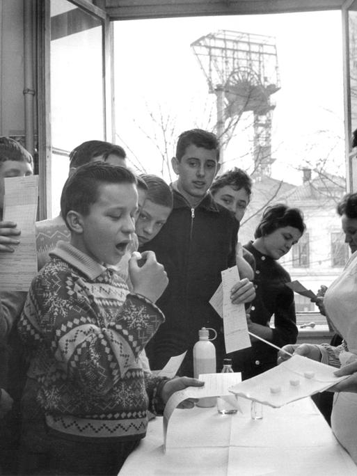 Eine Schwarz-Weißaufnahmen von 1962 zeigt Schulkinder, die unter Aufsicht einer Krankenschwester mit Impfstoff gegen Polio präparierte Zuckerwürfel einnehmen. 