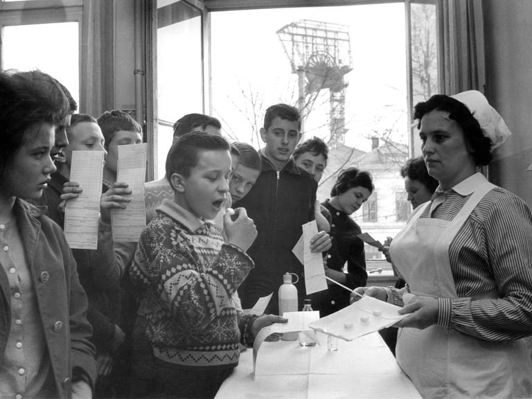 Eine Schwarz-Weißaufnahmen von 1962 zeigt Schulkinder, die unter Aufsicht einer Krankenschwester mit Impfstoff gegen Polio präparierte Zuckerwürfel einnehmen. 