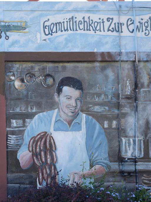Ein Wandbild im texanischen New Braunfels wirbt für ein deutsches Restaurant. Das Bild zeigt einen Koch, der Würstchen zubereitet, darüber der Schriftzug: Gemütlichkeit zur Ewigkeit. 