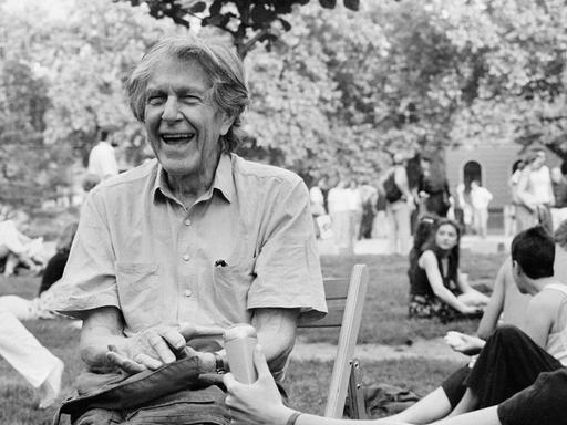 John Cage sitzt lachend auf einer Wiese und gibt ein Interview in ein Reportermikrofon, im Hintergrund sitzen und gehen junge Leute in Freizeitkleidung.