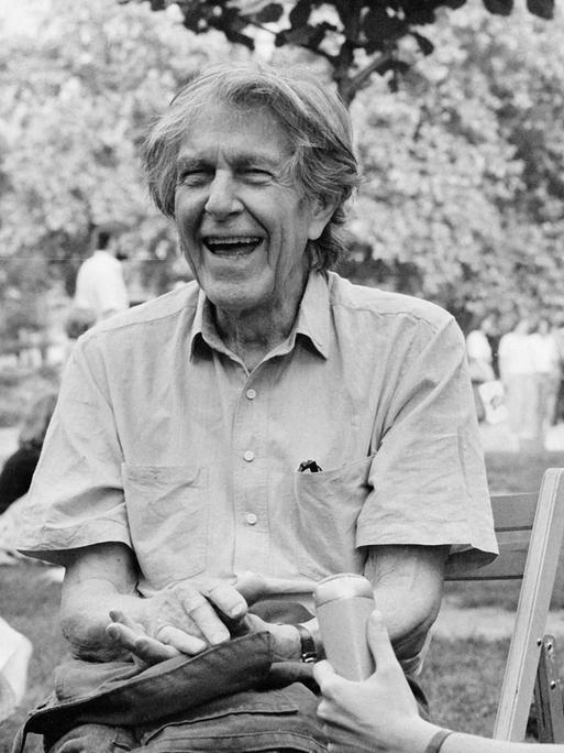 John Cage sitzt lachend auf einer Wiese und gibt ein Interview in ein Reportermikrofon, im Hintergrund sitzen und gehen junge Leute in Freizeitkleidung.