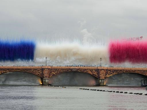 Die Pont d'Austerlitz über der Seine in Paris mit goldenen Applikationen. Darüber zeigen während der Eröffnungsfeier Rauchbomben die französische Nationalflagge.