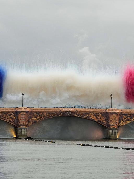 Die Pont d'Austerlitz über der Seine in Paris mit goldenen Applikationen. Darüber zeigen während der Eröffnungsfeier Rauchbomben die französische Nationalflagge.