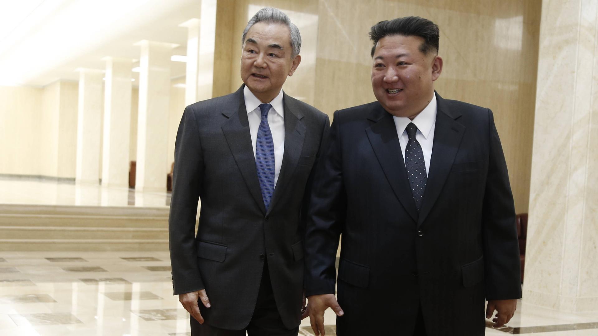 Nordkoreas Machthaber Kim Jong Un und der chinesische Außenminister Wang Yi stehe eng nebeneinander in einem großen Raum oder Gang mit hellem Marmor. 