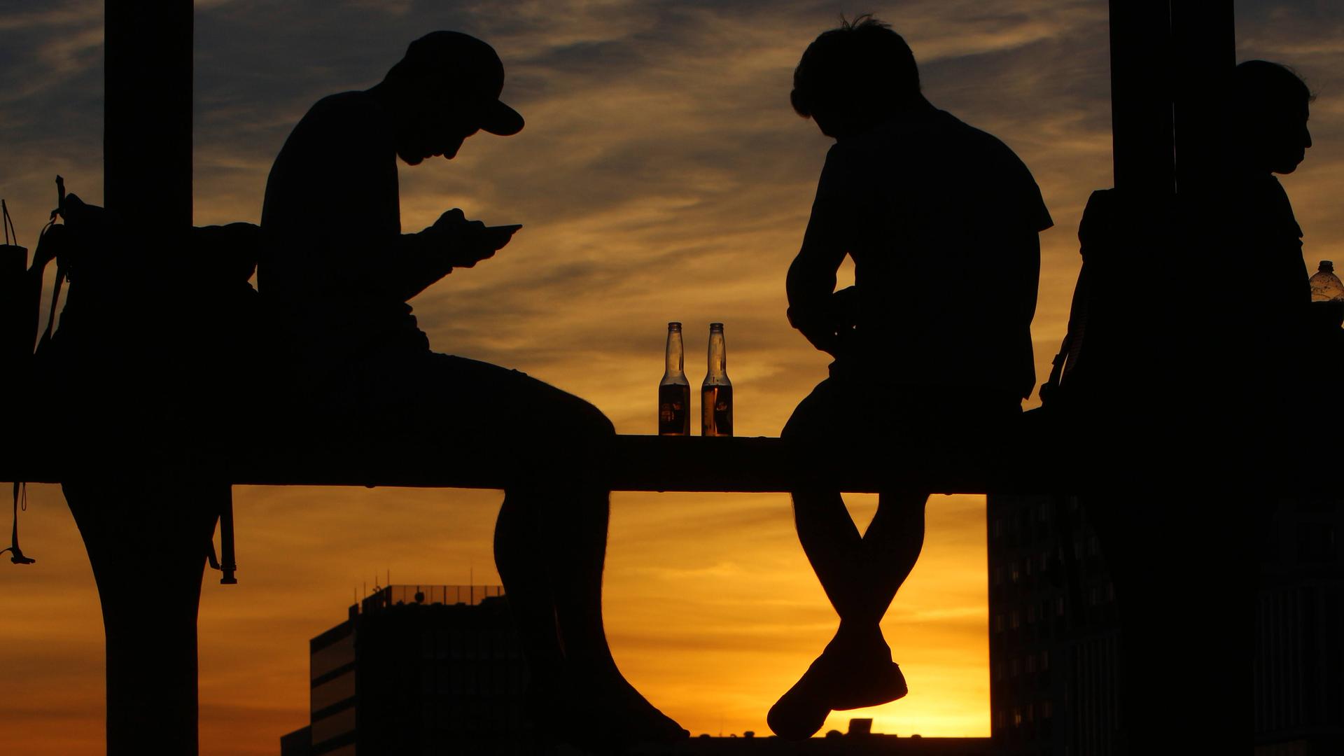 Junge Menschen sitzen auf dem Geländer einer Brücke vor dem Sonnenuntergang und blicken auf ihr Smartphone. Junge Menschen sitzen auf dem Geländer einer Brücke vor dem Sonnenuntergang und blicken auf ihr Smartphone.
