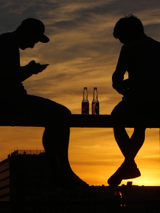 Junge Menschen sitzen auf dem Geländer einer Brücke vor dem Sonnenuntergang und blicken auf ihr Smartphone. Junge Menschen sitzen auf dem Geländer einer Brücke vor dem Sonnenuntergang und blicken auf ihr Smartphone.
