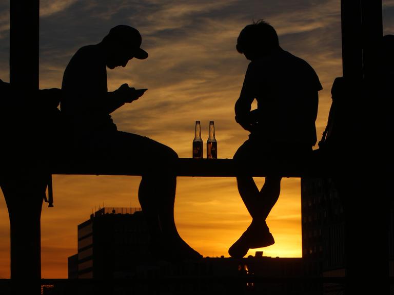 Junge Menschen sitzen auf dem Geländer einer Brücke vor dem Sonnenuntergang und blicken auf ihr Smartphone. Junge Menschen sitzen auf dem Geländer einer Brücke vor dem Sonnenuntergang und blicken auf ihr Smartphone.