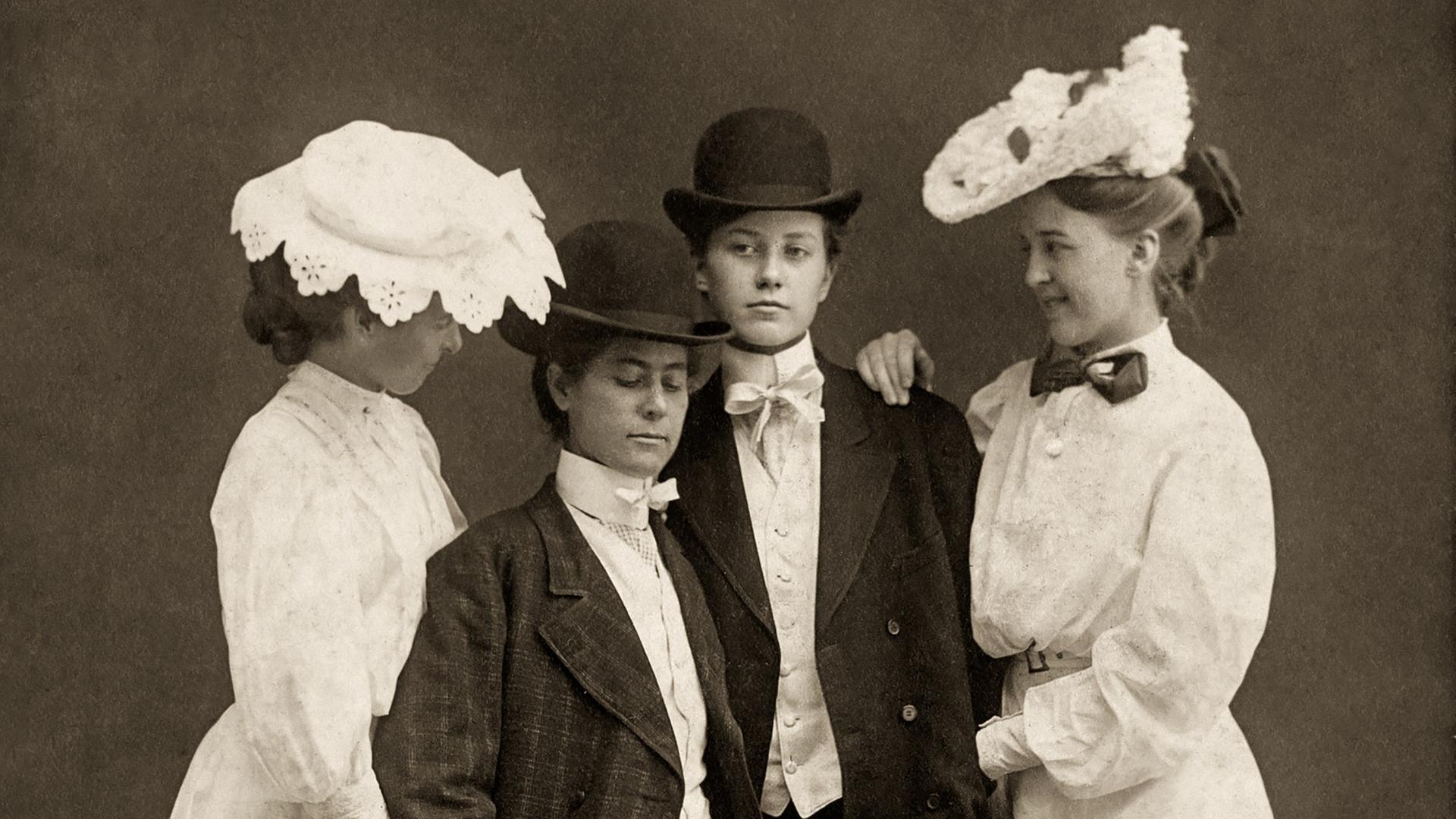 Auf einer historischen Aufnahme sind vier Frauen zu sehen: Zwei von ihnen tragen lange Kleider, zwei Anzug und eine Melone auf dem Kopf. Auf einer historischen Aufnahme sind vier Frauen zu sehen: Zwei von ihnen tragen lange Kleider, zwei Anzug und eine Melone auf dem Kopf.
