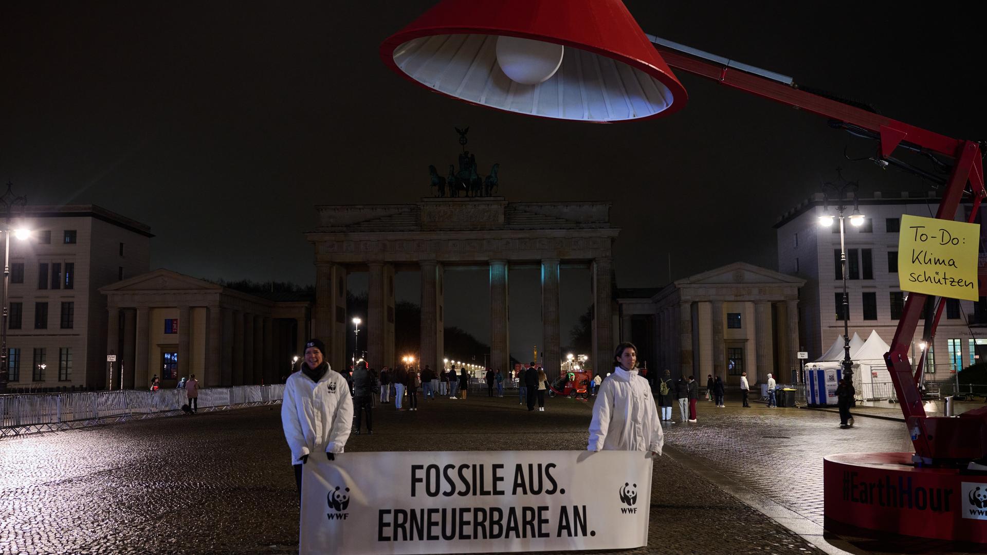 „Fossile aus. Erneuerbare an.“ steht auf einem Banner unter einer überdimensionalen Lampe vor dem dunklen Brandenburger Tor. 