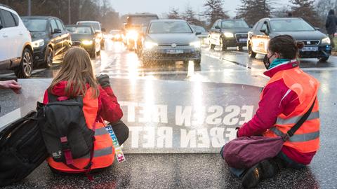 Zwei junge Frauen in orangen Warnwesten mit einem Transparent knien auf einer Fahrbahn, um sie zu blockieren. Ihnen gegenüber: PKWs mit leuchtenden Scheinwerfern. 