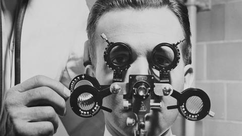Eine historische schwarz-weiß Fotografie zeigt den Kopf eines Mannes hinter einem Augenmessgerät in einen Klinik.