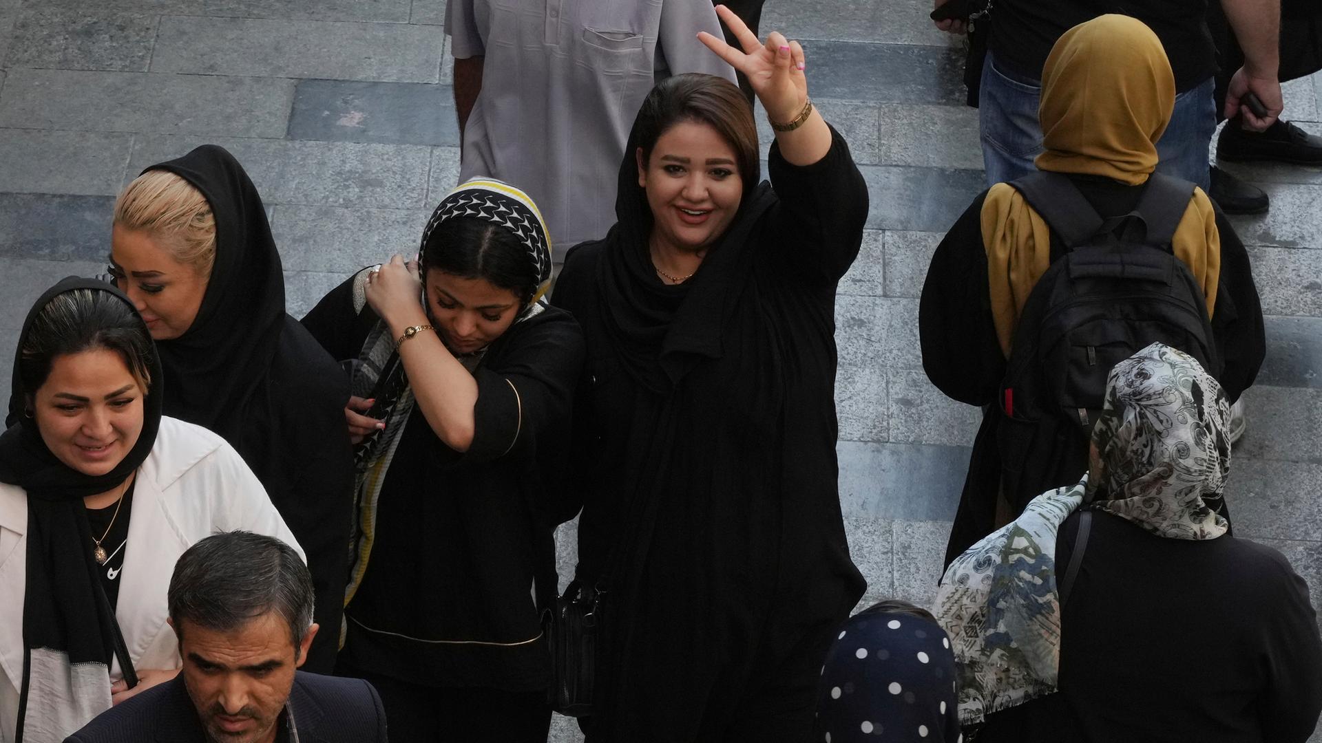 Eine breit lächelnde Frau in Teheran macht das Victory-Zeichen, sie trägt die Haare offen. Einige Frauen neben ihr tragen ein Kopftuch.