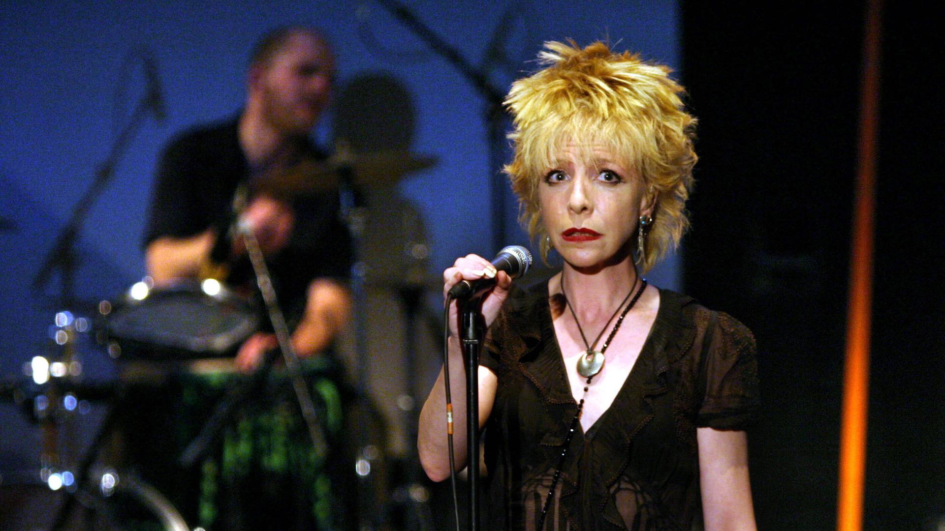 Die Sängerin Julee Cruise steht in der Berliner Volksbühne hinter einem Mikro und guckt unfroh. Im Hintergrund steht ein Schlagzeug.