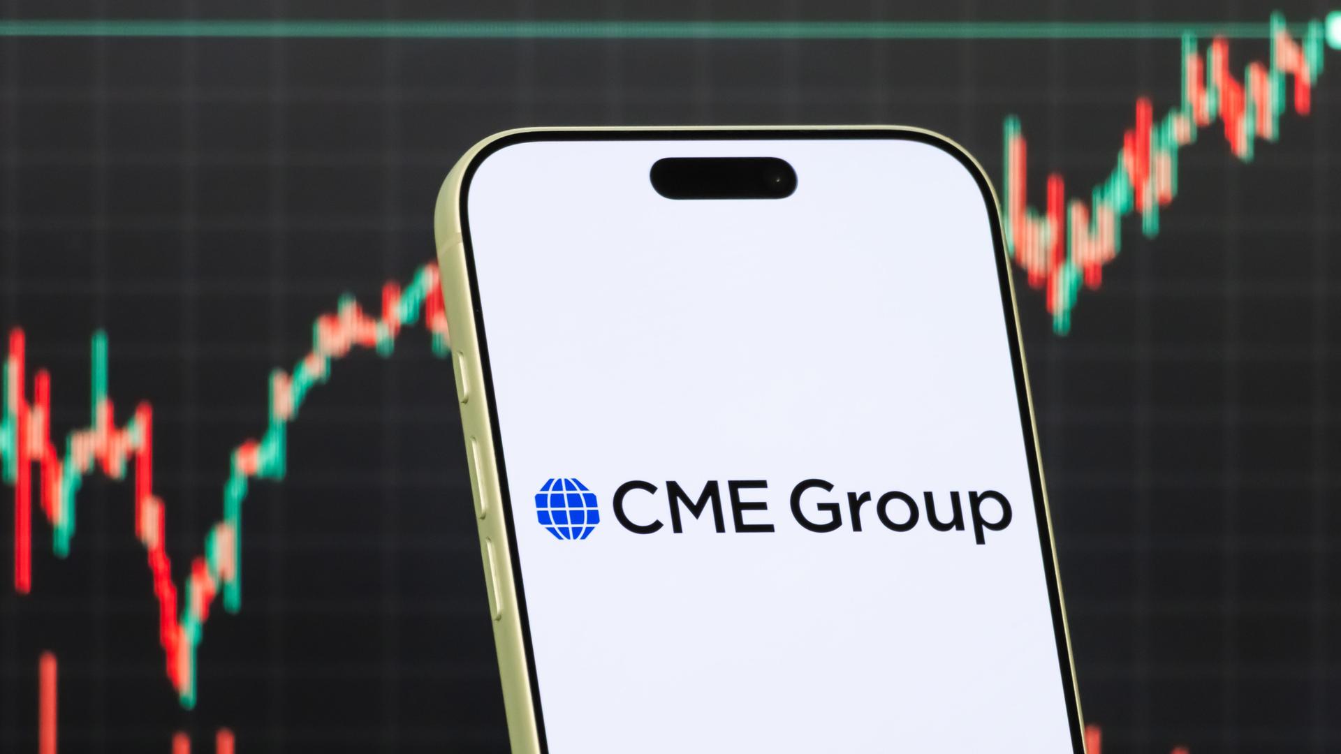 Diese Fotomontage zeigt das Logo der CME Group auf einem Smartphone vor einem Bildschirm, der den aktuellen Aktienmarkt-Chart des Unternehmens anzeigt. 