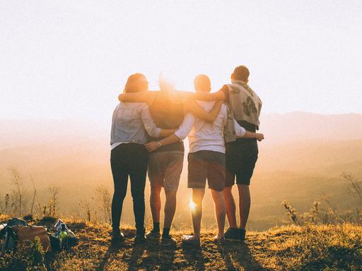 Vier Personen liegen sich in den Armen und schauen gemeinsam in einen Sonnenuntergang.