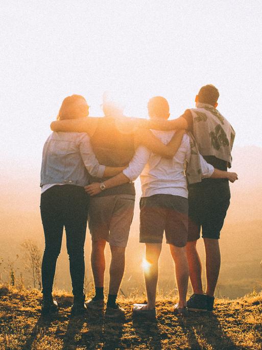 Vier Personen liegen sich in den Armen und schauen gemeinsam in einen Sonnenuntergang.