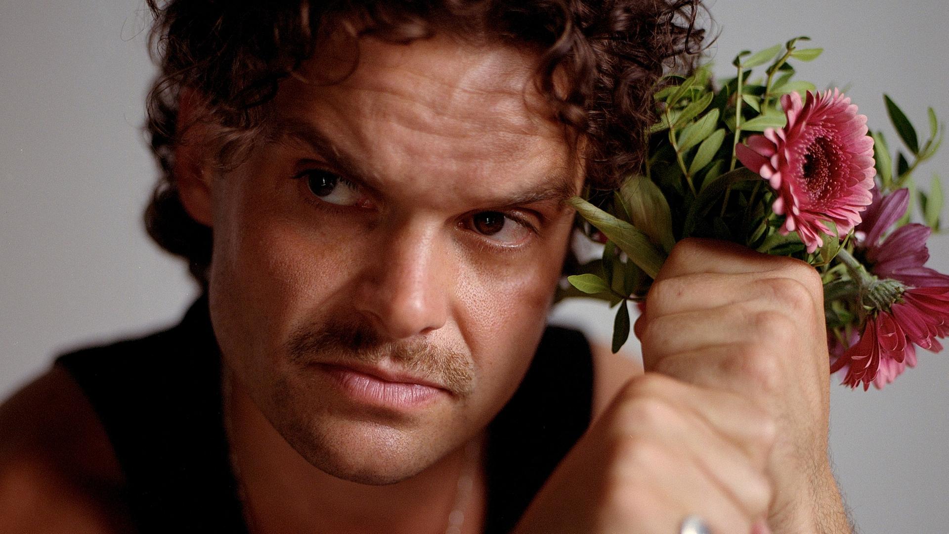 Ein Mann mit braunen Locken und Schnauzbart schaut ernst aus dem Bild hinaus. Neben dem Kopf hält er in seinen Fäusten einen Blumenstrauß mit rosa Gerbera. Er trägt ein schwarzes Tanktop und silbernen Schmuck. Ein Mann mit braunen Locken und Schnauzbart schaut ernst aus dem Bild hinaus. Neben dem Kopf hält er in seinen Fäusten einen Blumenstrauß mit rosa Gerbera. Er trägt ein schwarzes Tanktop und silbernen Schmuck.