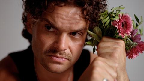 Ein Mann mit braunen Locken und Schnauzbart schaut ernst aus dem Bild hinaus. Neben dem Kopf hält er in seinen Fäusten einen Blumenstrauß mit rosa Gerbera. Er trägt ein schwarzes Tanktop und silbernen Schmuck.