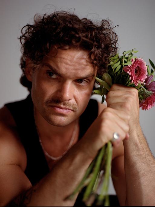 Ein Mann mit braunen Locken und Schnauzbart schaut ernst aus dem Bild hinaus. Neben dem Kopf hält er in seinen Fäusten einen Blumenstrauß mit rosa Gerbera. Er trägt ein schwarzes Tanktop und silbernen Schmuck.
