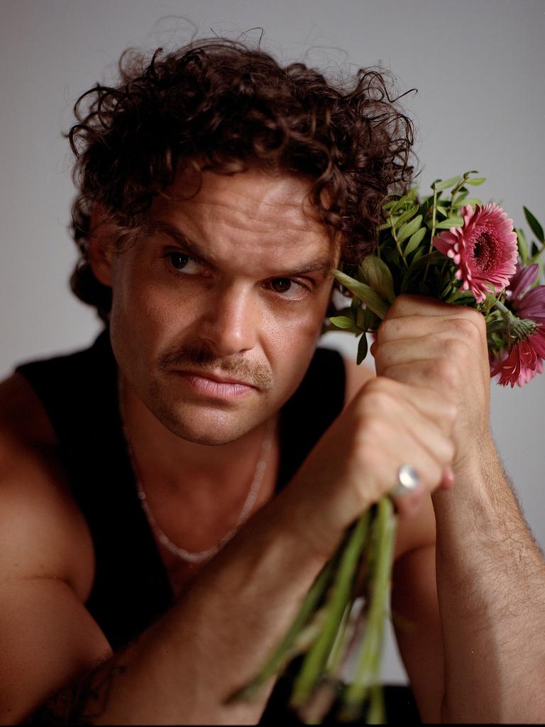 Ein Mann mit braunen Locken und Schnauzbart schaut ernst aus dem Bild hinaus. Neben dem Kopf hält er in seinen Fäusten einen Blumenstrauß mit rosa Gerbera. Er trägt ein schwarzes Tanktop und silbernen Schmuck.