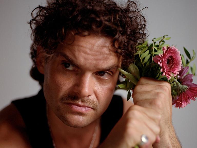 Ein Mann mit braunen Locken und Schnauzbart schaut ernst aus dem Bild hinaus. Neben dem Kopf hält er in seinen Fäusten einen Blumenstrauß mit rosa Gerbera. Er trägt ein schwarzes Tanktop und silbernen Schmuck.