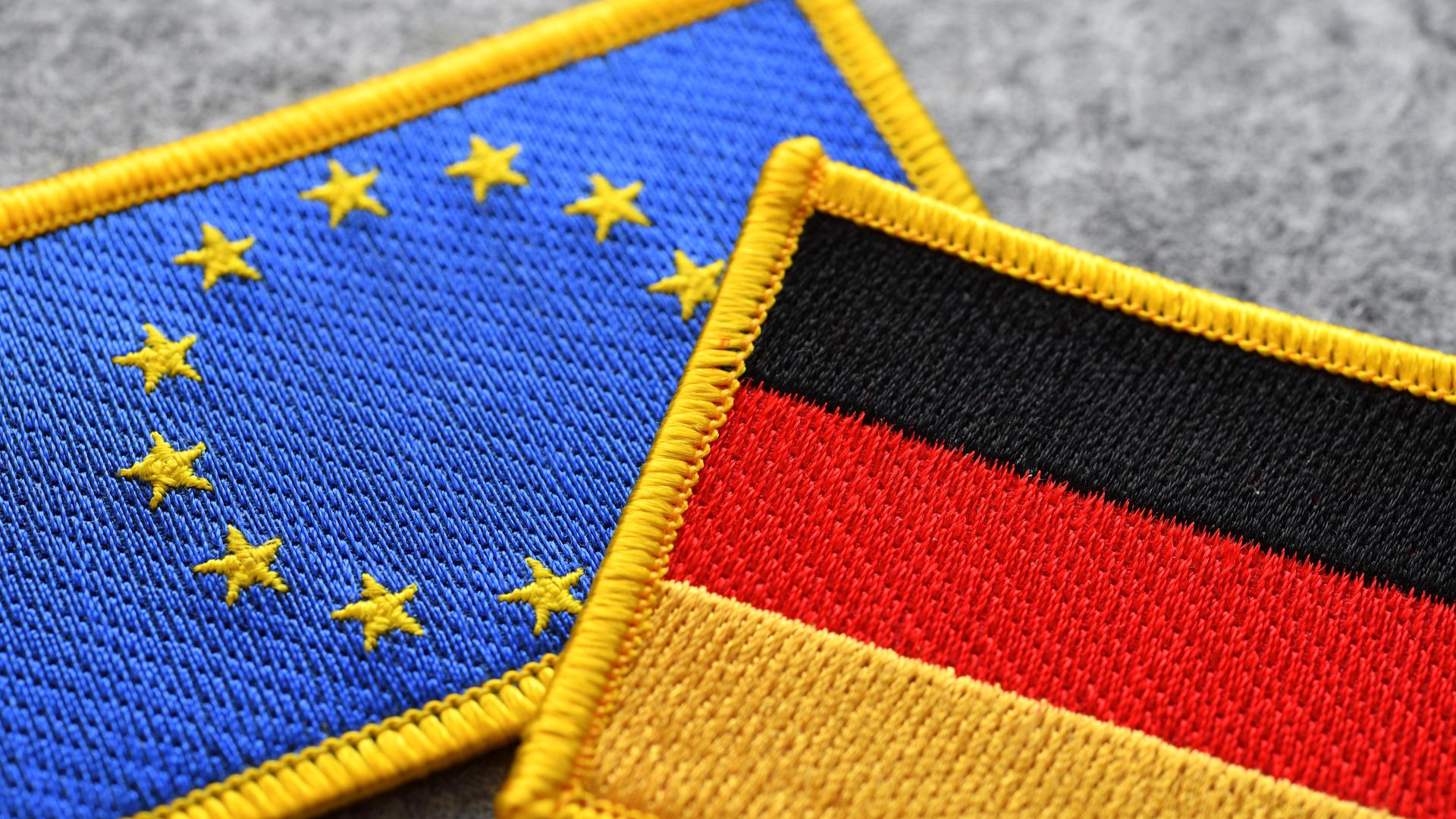Zwei Aufnäher, einer mit der europäischen Flagge, der andere mit der deutschen Flagge, liegen auf einem grauen Untergrund. Zwei Aufnäher, einer mit der europäischen Flagge, der andere mit der deutschen Flagge, liegen auf einem grauen Untergrund.