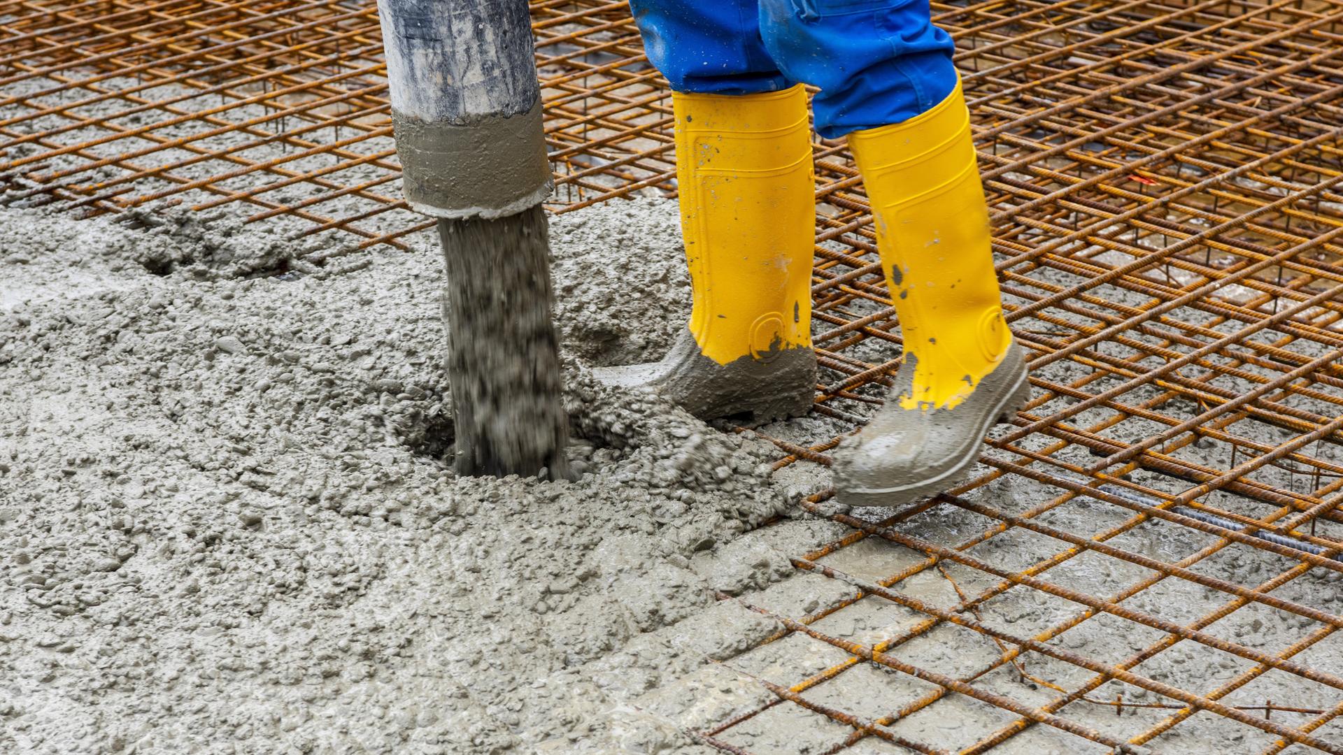 Zu sehen sind die Füße in gelben Gummistiefeln eines Bauarbeiters, der Zement auf einem Boden verteilt