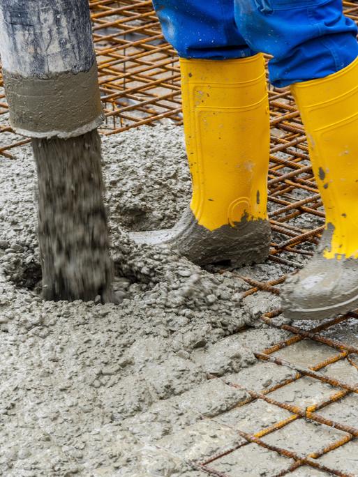 Zu sehen sind die Füße in gelben Gummistiefeln eines Bauarbeiters, der Zement auf einem Boden verteilt