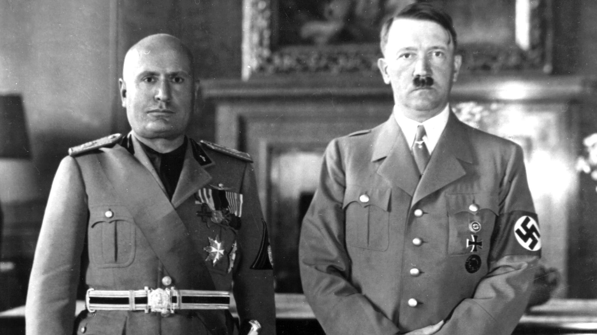 Hitler und Mussolini stehen nebeneinander. Sie tragen Uniform und schauen streng in die Kamera.