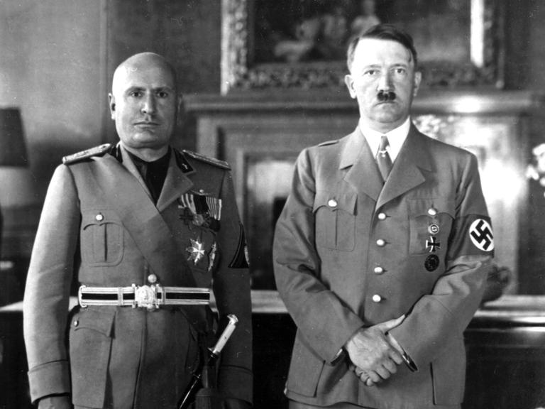 Hitler und Mussolini stehen nebeneinander. Sie tragen Uniform und schauen streng in die Kamera.