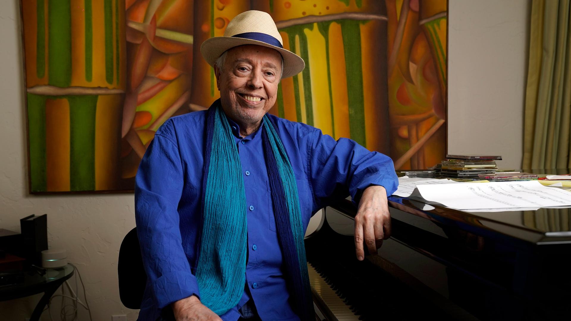 Sergio Mendes sitzt mit Hut an einem Flügel, im Hintergrund ein buntes Gemälde. Er lacht in die Kamer. 